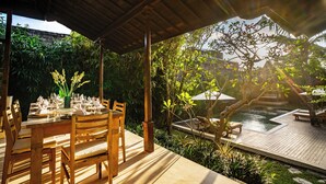 Un área de comedor al aire libre con una mesa de madera lista para una comida, rodeada de abundante vegetación y una pequeña piscina.