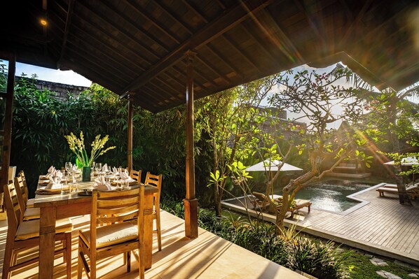 An outdoor dining area with a wooden table set for a meal, surrounded by lush greenery and a small pool.