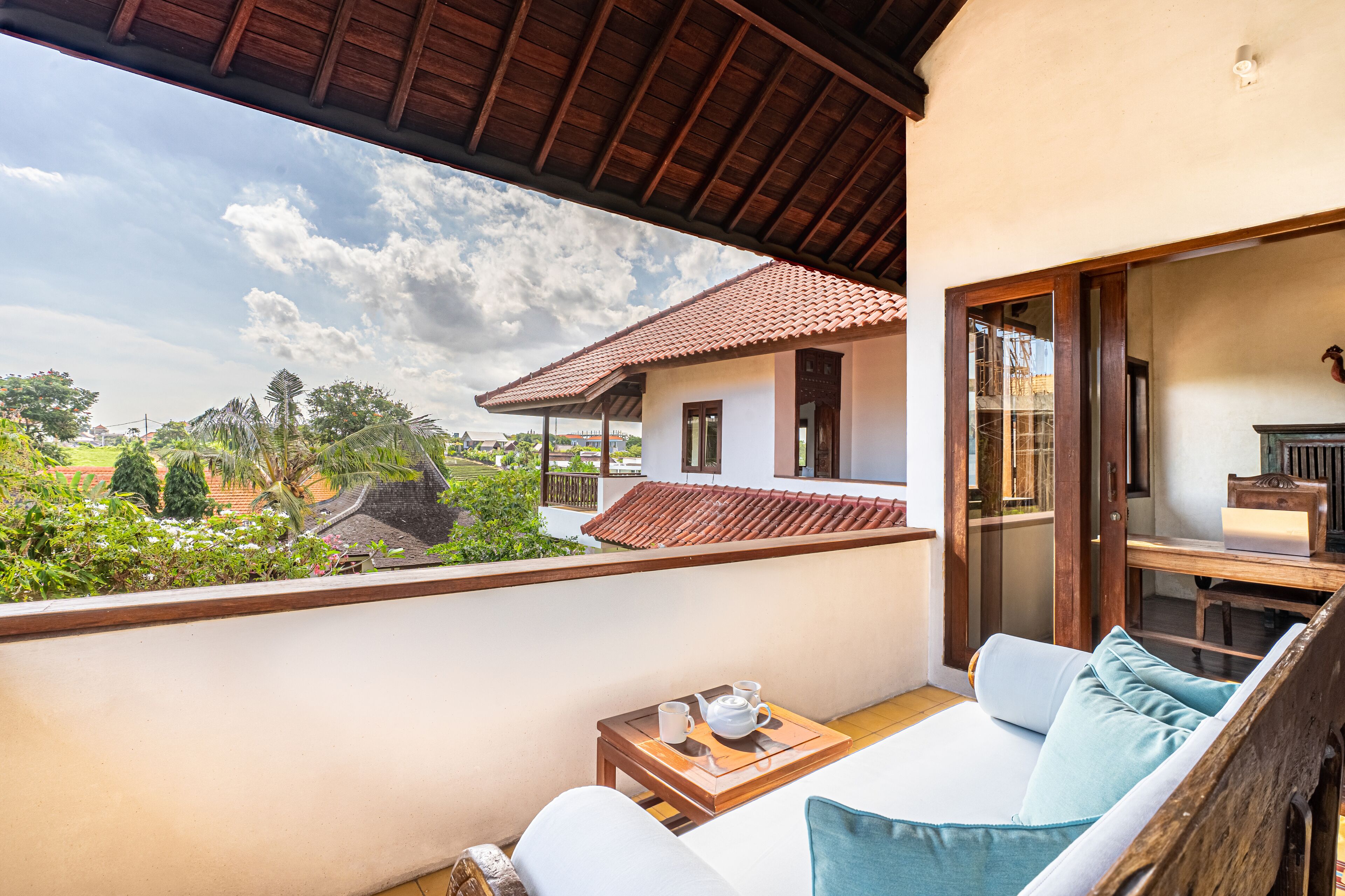 Ein Balkon mit Blick auf ein Haus und einen Garten, eine Sitzecke mit Kissen und ein kleiner Tisch mit Teekanne und Tassen.