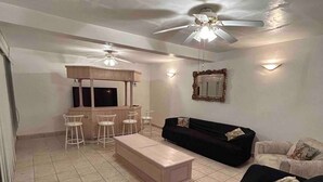 A living room with a black sofa, a wooden coffee table, and a TV stand with a flat-screen TV. There are ceiling fans, a mirror, and a tiled floor. - Charming 4-bedroom house with WiFi in Playas de Rosarito, secure gated parking (Playas de Rosarito)