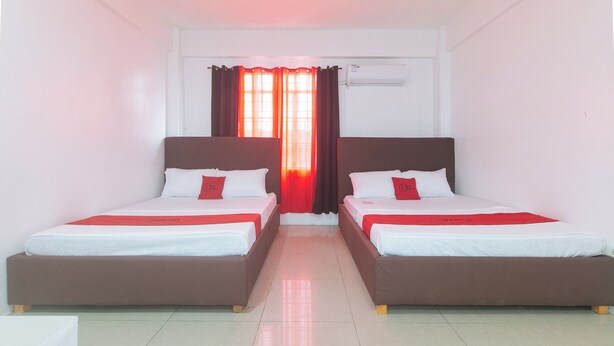 Een hotelkamer met twee eenpersoonsbedden, elk met een rood accentkussen, witte beddengoed en een bruin hoofdeinde. Een raam met rode gordijnen laat natuurlijk licht de kamer binnen.