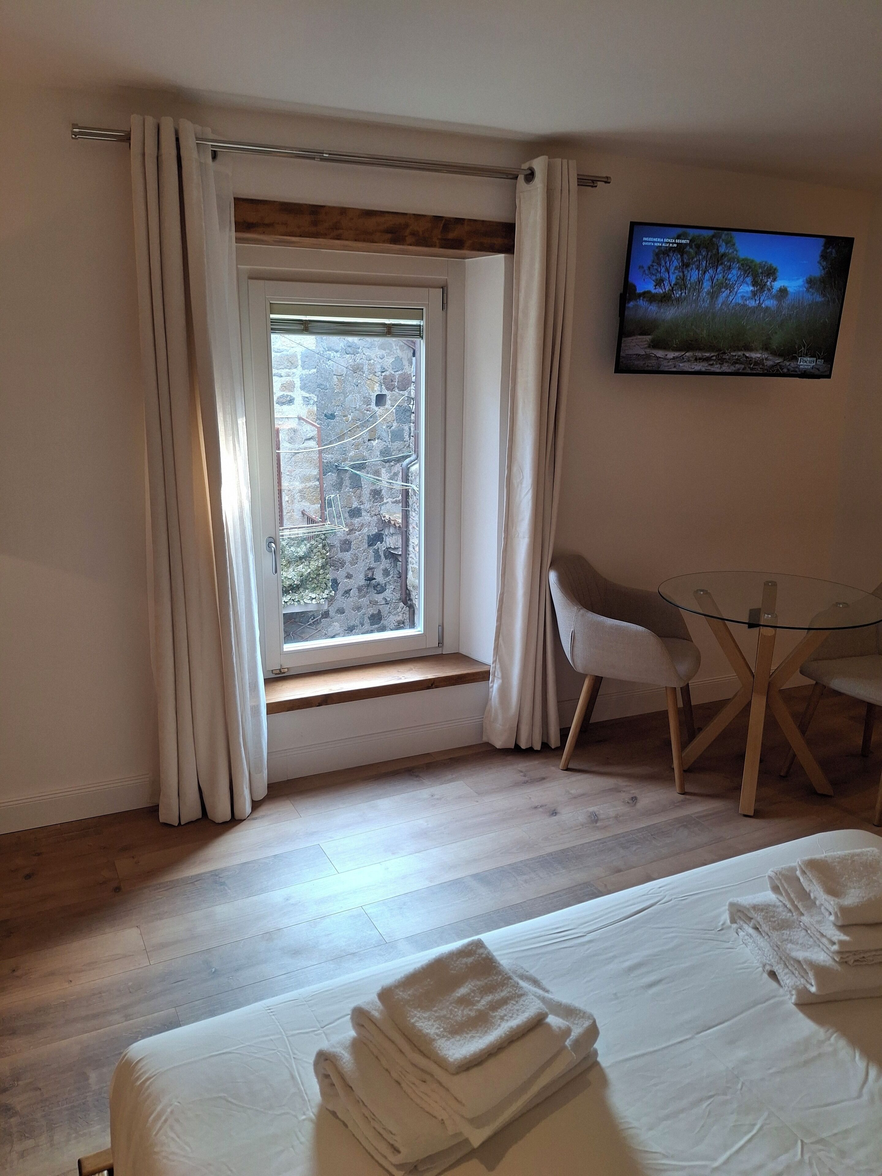 Une chambre d’hôtel avec une grande fenêtre, un lit avec des serviettes soigneusement pliées, une table en verre avec des chaises et un téléviseur fixé au mur.
