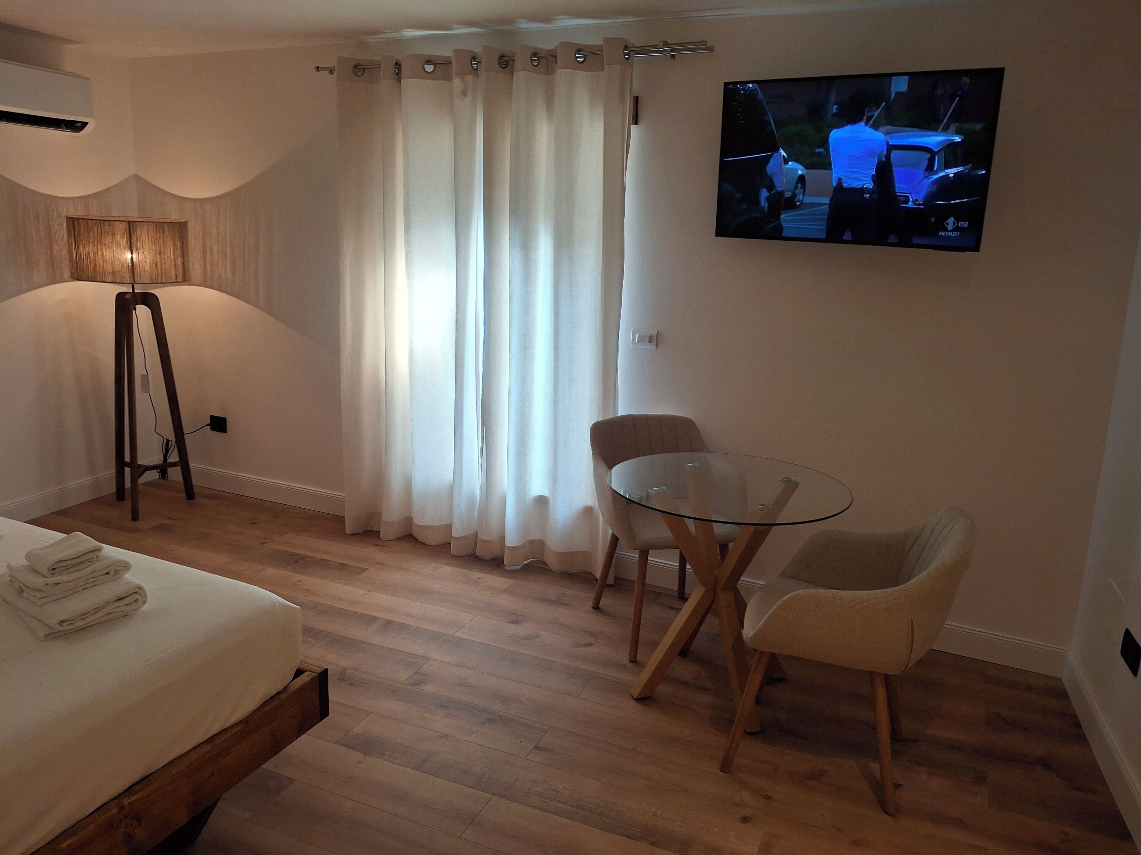 A hotel room with a bed, a glass table with two chairs, a TV on the wall, and a floor lamp.