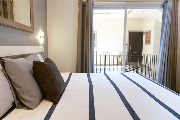 A hotel room with a bed, pillows, a window with curtains, and a view of a pool and balcony.