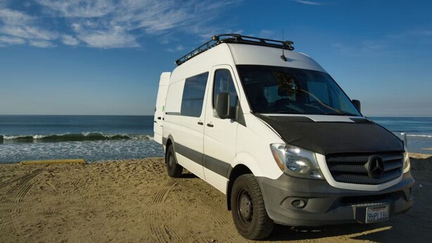 A white van parked on a sandy beach with ocean waves in the background.