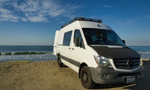 A white van parked on a sandy beach with ocean waves in the background.