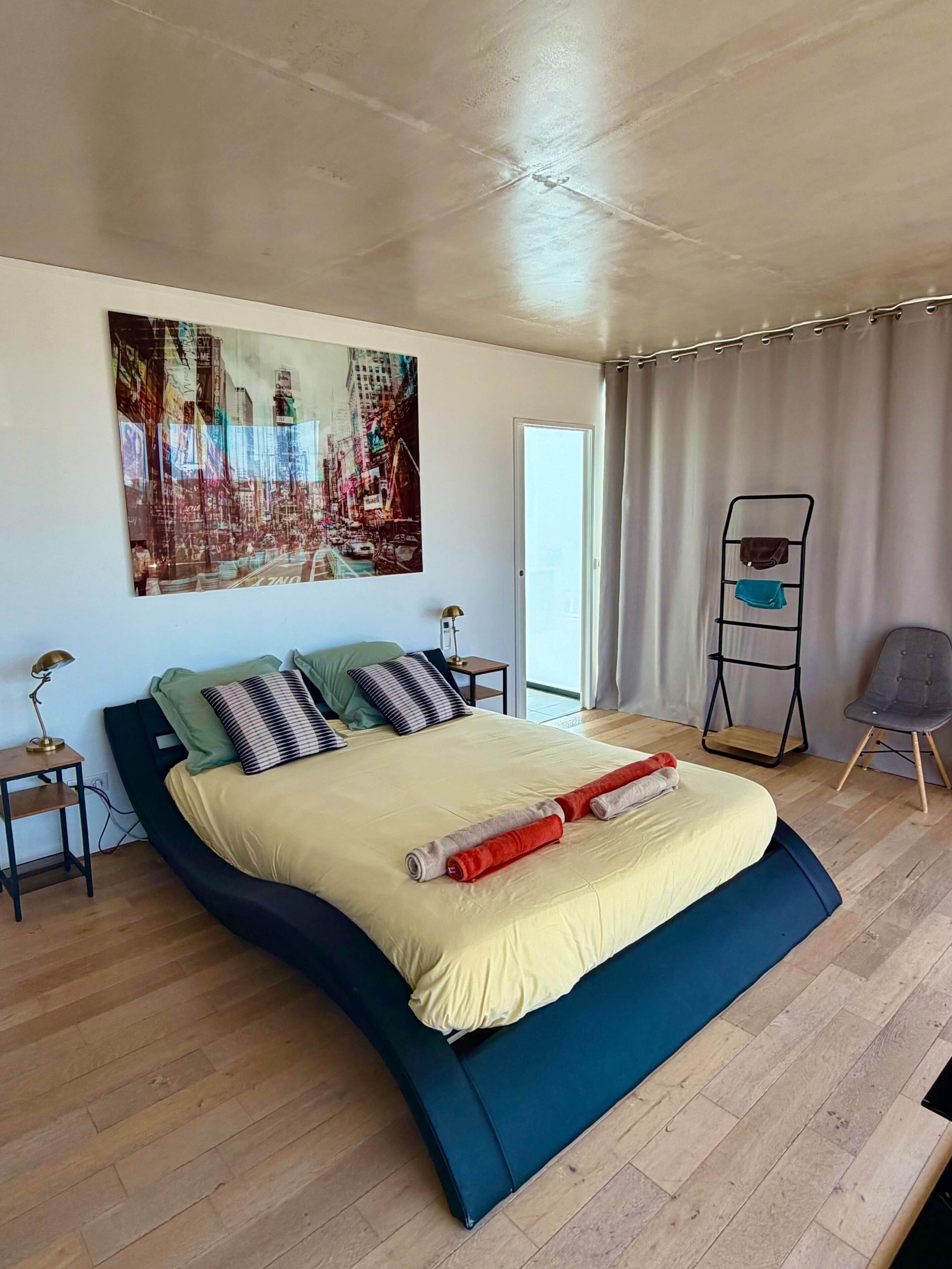 A modern bedroom with a large bed, a nightstand, a lamp, a chair, a small table, and a painting on the wall.