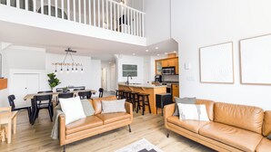 A spacious living area with a high ceiling, a dining table, two leather sofas, and a kitchen in the background.