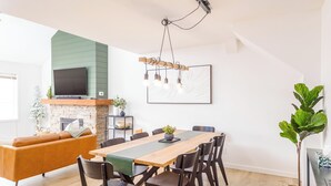 A modern living room with a wooden dining table, dark chairs, a sofa, a fireplace, and a flat-screen TV mounted on the wall.