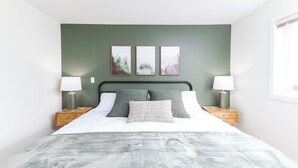 A neatly made bed with a grey bedspread and two pillows, flanked by two bedside tables with lamps, in a room with green walls and three framed pictures above the bed.