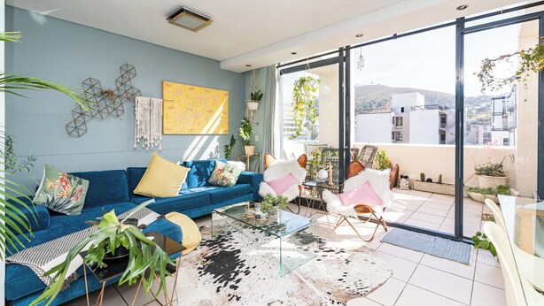 A modern living room with a blue sofa, a glass coffee table, and a large window overlooking a cityscape. - Cape Eazi Stayz The Decks (Cape Town)