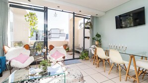 A modern living room with a glass coffee table, plush chairs, a glass dining table with chairs, and a TV mounted on the wall. - Cape Eazi Stayz The Decks (Cape Town)