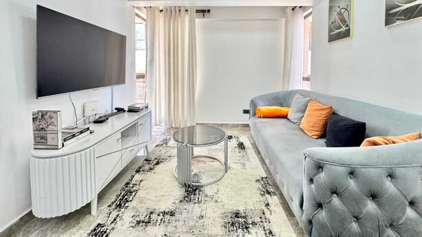 Una sala de estar moderna con un televisor de pantalla plana, un sofá gris con cojines anaranjados, un mueble blanco para entretenimiento y una mesa de centro redonda.