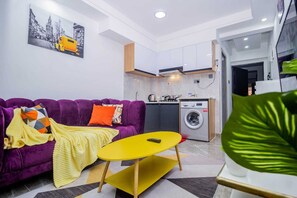 A compact living space with a purple sofa, a yellow table, a washing machine, and a kitchen area.