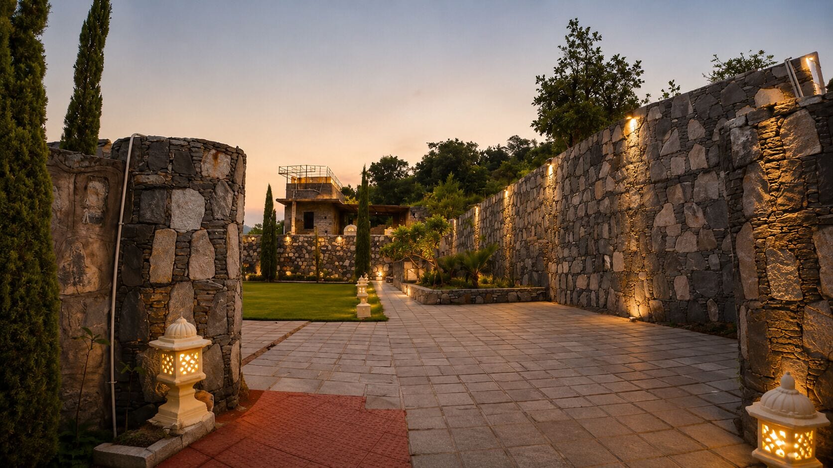 Un sendero de piedra que lleva a un edificio con fachada de piedra, rodeado de follaje e iluminado por luces empotradas.