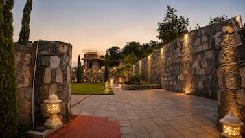 Un sendero de piedra que lleva a un edificio con fachada de piedra, rodeado de follaje e iluminado por luces empotradas.