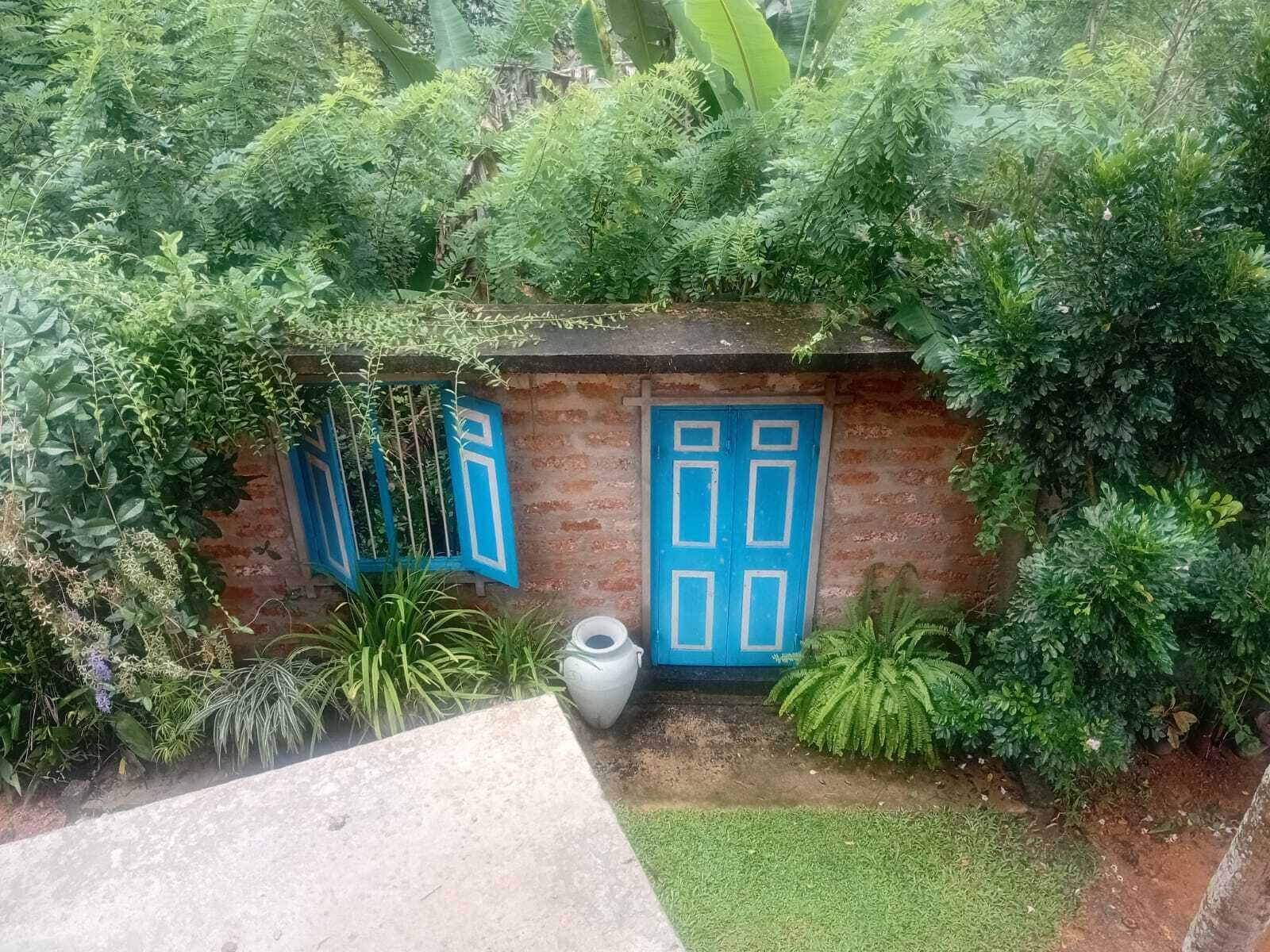 Una pequeña construcción de ladrillo con puertas y ventanas azules, rodeada de abundante vegetación.