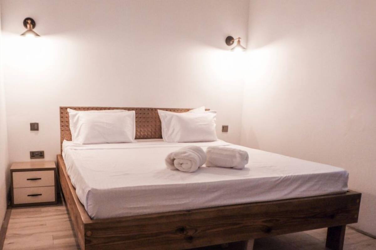 Een net opgemaakt bed met witte lakens en opgerolde handdoeken, een houten hoofdbord, en een nachtkastje met lades.