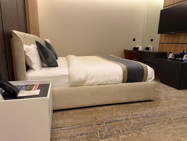 A hotel room with a bed, a desk with a phone and a magazine, a TV, and a minibar. - White Season Hotel (Haql)
