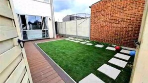 Een achtertuin met een houten terras, een gazon en een bakstenen muur.