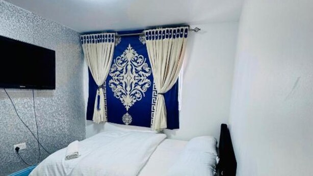 A bedroom with a bed, curtains, a mounted TV, and a bedside table with a speaker.