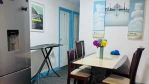 A compact kitchen with a refrigerator, a dining table with chairs, a water dispenser, and a small table with a vase of flowers.