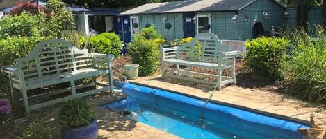 A pool area with two white benches, a small pool, and a shed in the background.