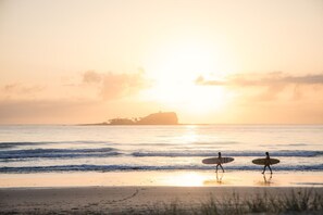 Two people walking on a beach with surfboards, a sunset in the background, and a distant landmass.