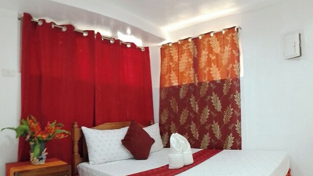 Una cama individual con una colcha roja y blanca, una mesita de noche de madera con un florero y cortinas rojas.
