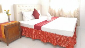 Una cama bien tendida con una colcha roja y blanca, una mesita de noche de madera con un jarrón de flores y una ventana con cortinas translúcidas.