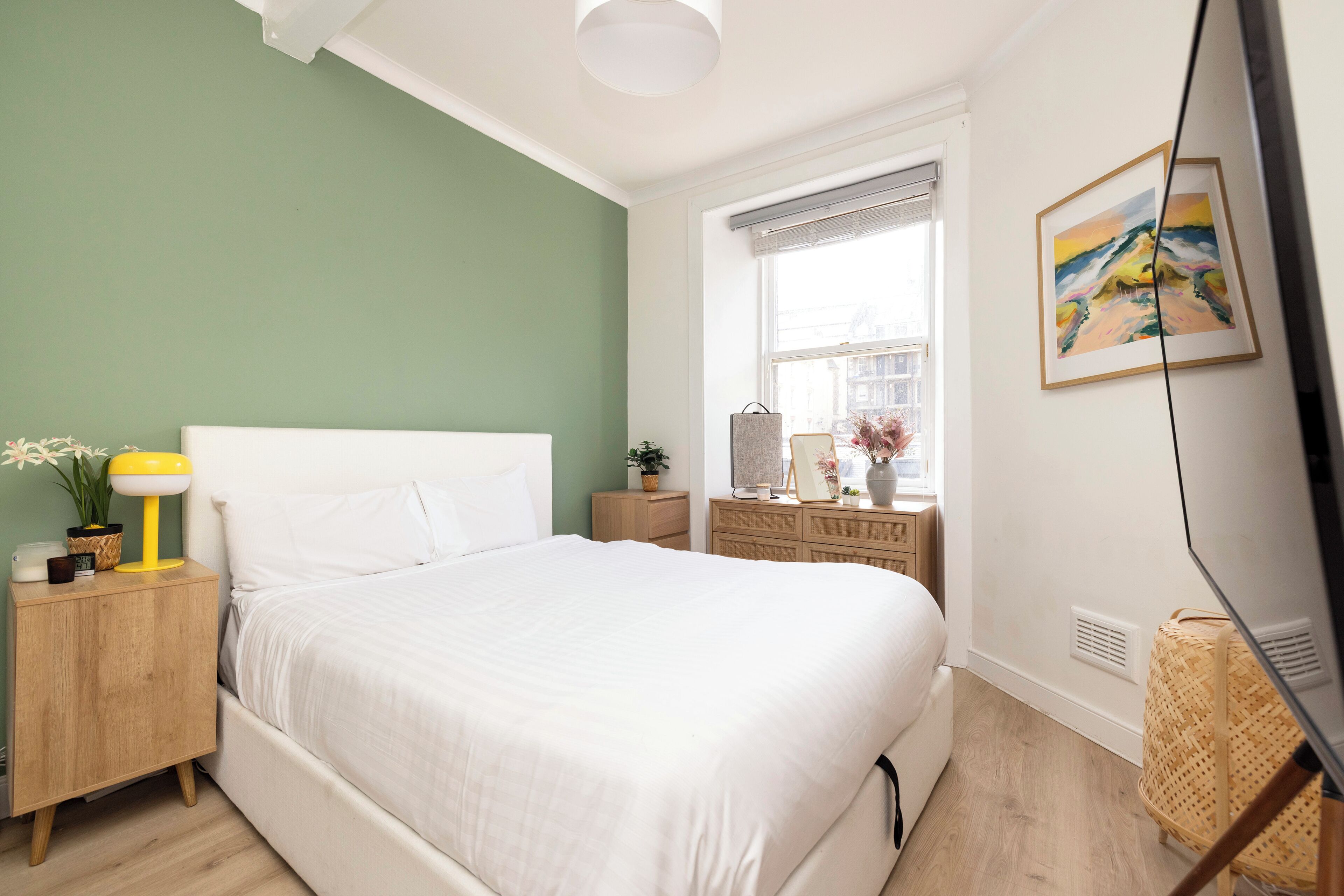 A bedroom with a bed, bedside table, lamp, dresser, and a painting on the wall.