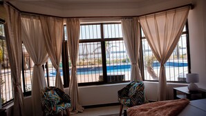 A hotel room with a large bay window, two patterned armchairs, a bed, and a nightstand with a lamp.