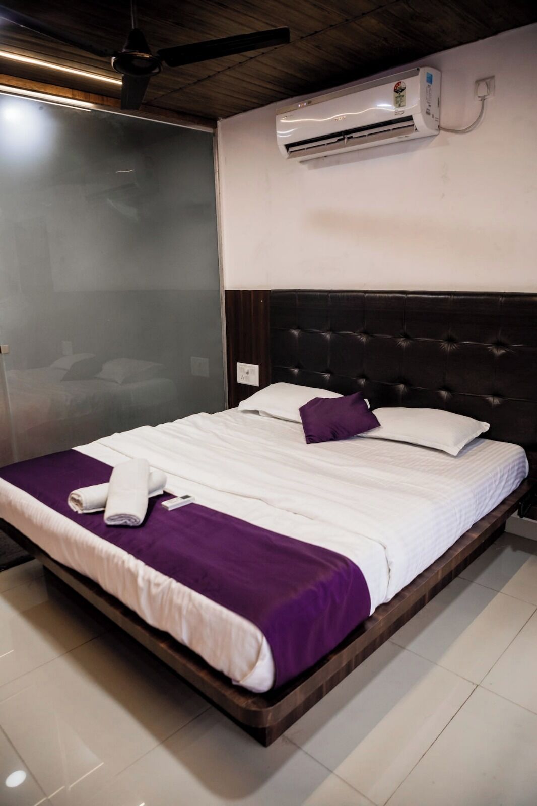 Una cama bien hecha con una colcha morada y sábanas blancas, un cabecero oscuro y un equipo de aire acondicionado instalado en la pared.