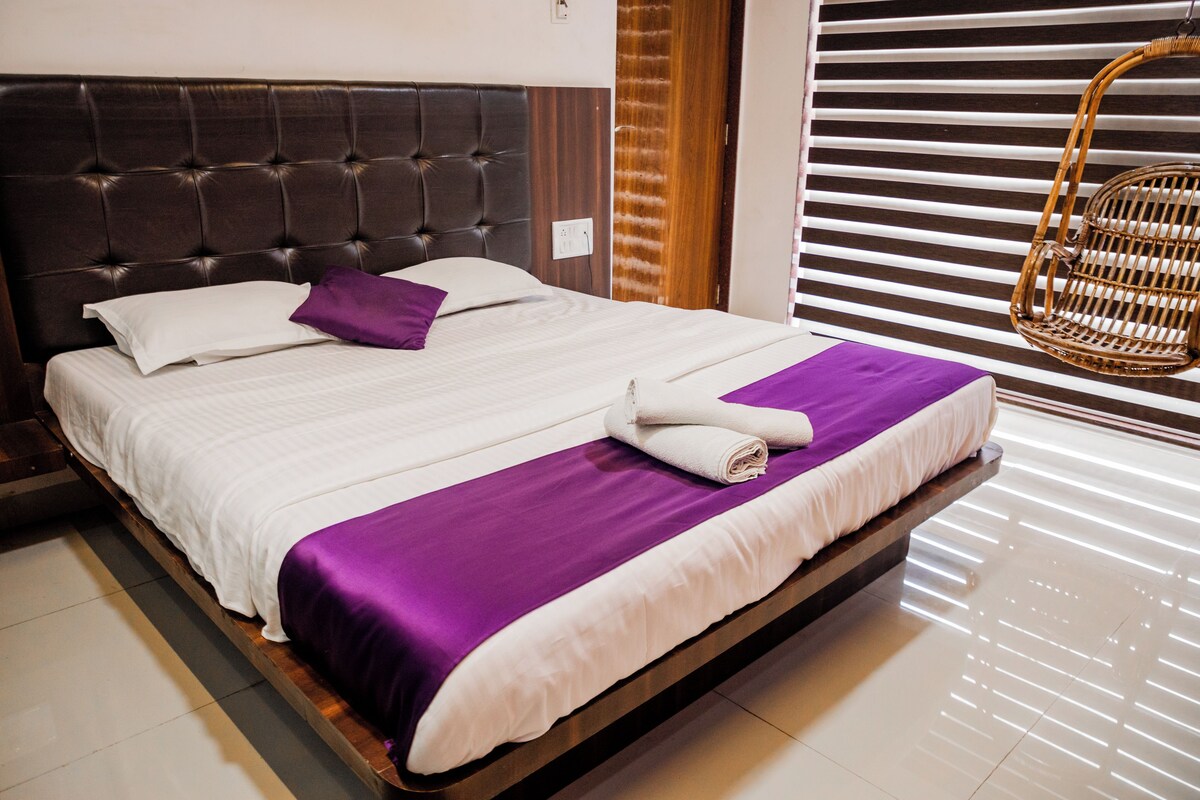 A neatly made bed with a white and purple bedspread, a wooden headboard, and a wicker chair in the corner.