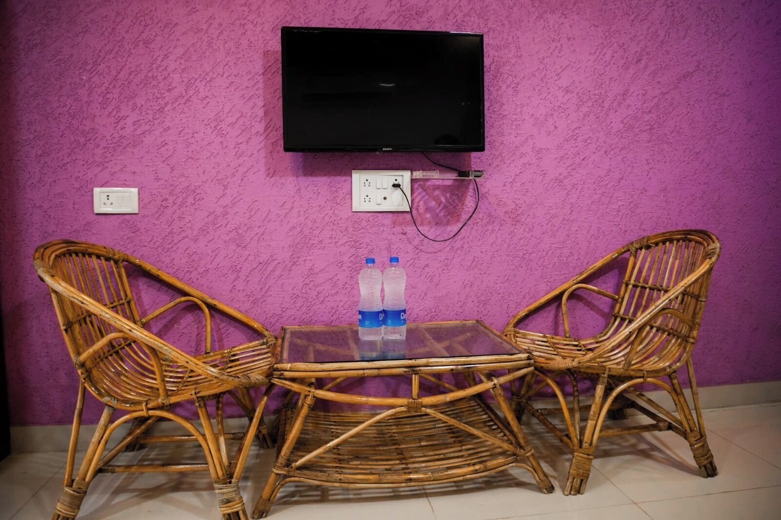 Une pièce avec un mur violet à la texture particulière, un téléviseur à écran plat fixé au-dessus d’une table, deux chaises en osier et une prise électrique sur le mur.