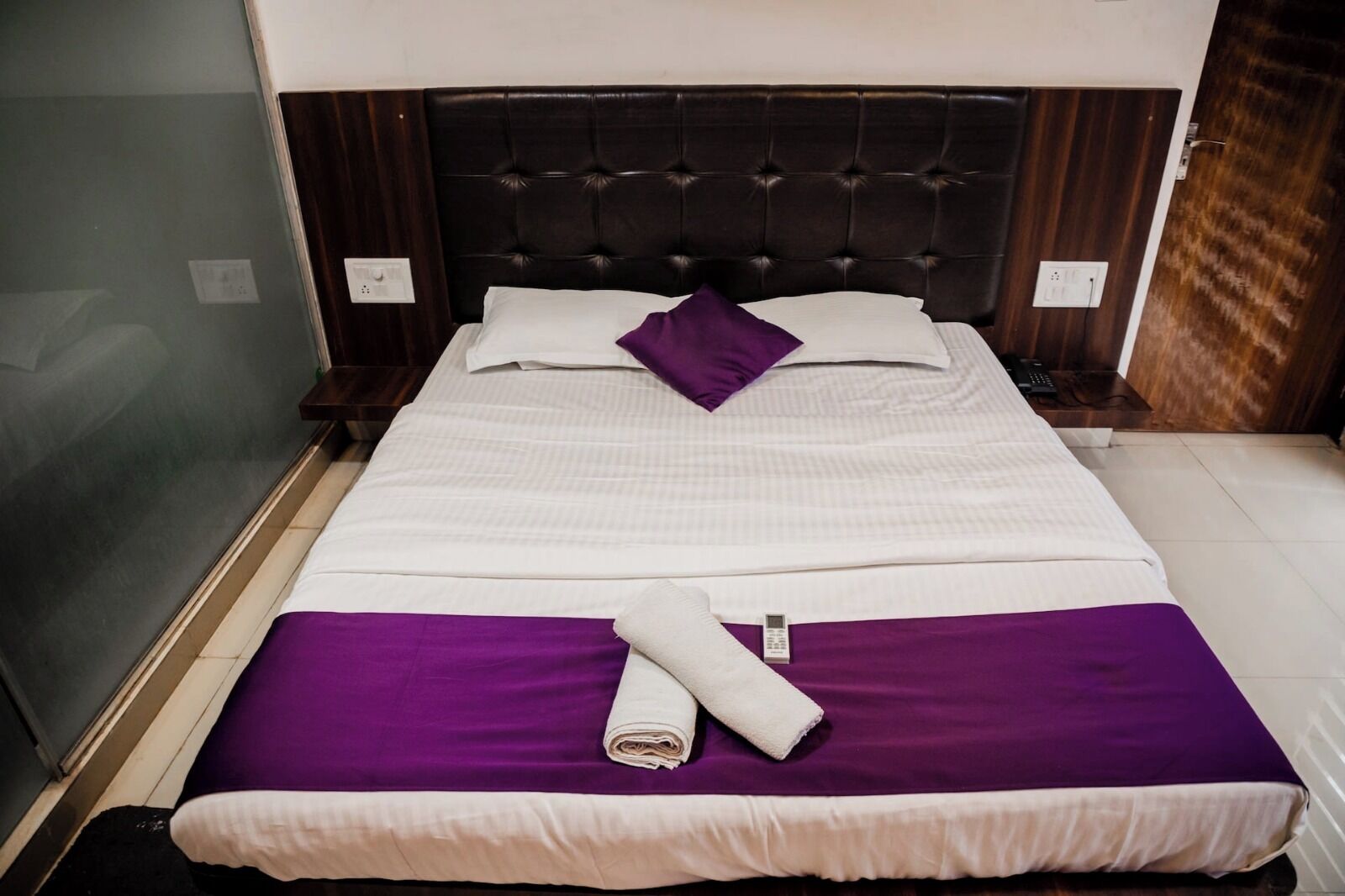 A neatly made bed with a purple and white bedspread, a rolled-up towel, and a remote control on the bed.