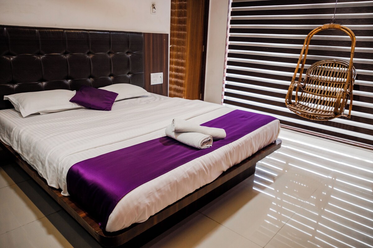 A neatly made bed with a purple blanket and white pillows, a wooden headboard, and a wicker chair by a window with blinds.