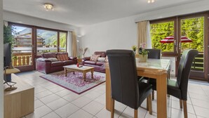 A spacious living room with a sofa set, a dining table with chairs, and a large window with a view of greenery. - View, 2 terraces & private parking - Saas-Grund (Saas-Grund)