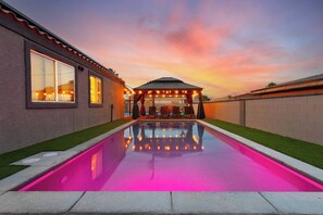 A swimming pool with a pink hue, a covered patio area, and a sunset sky. - 5BR Poolside Pickleball Retreat with Outdoor Oasis (North Las Vegas)