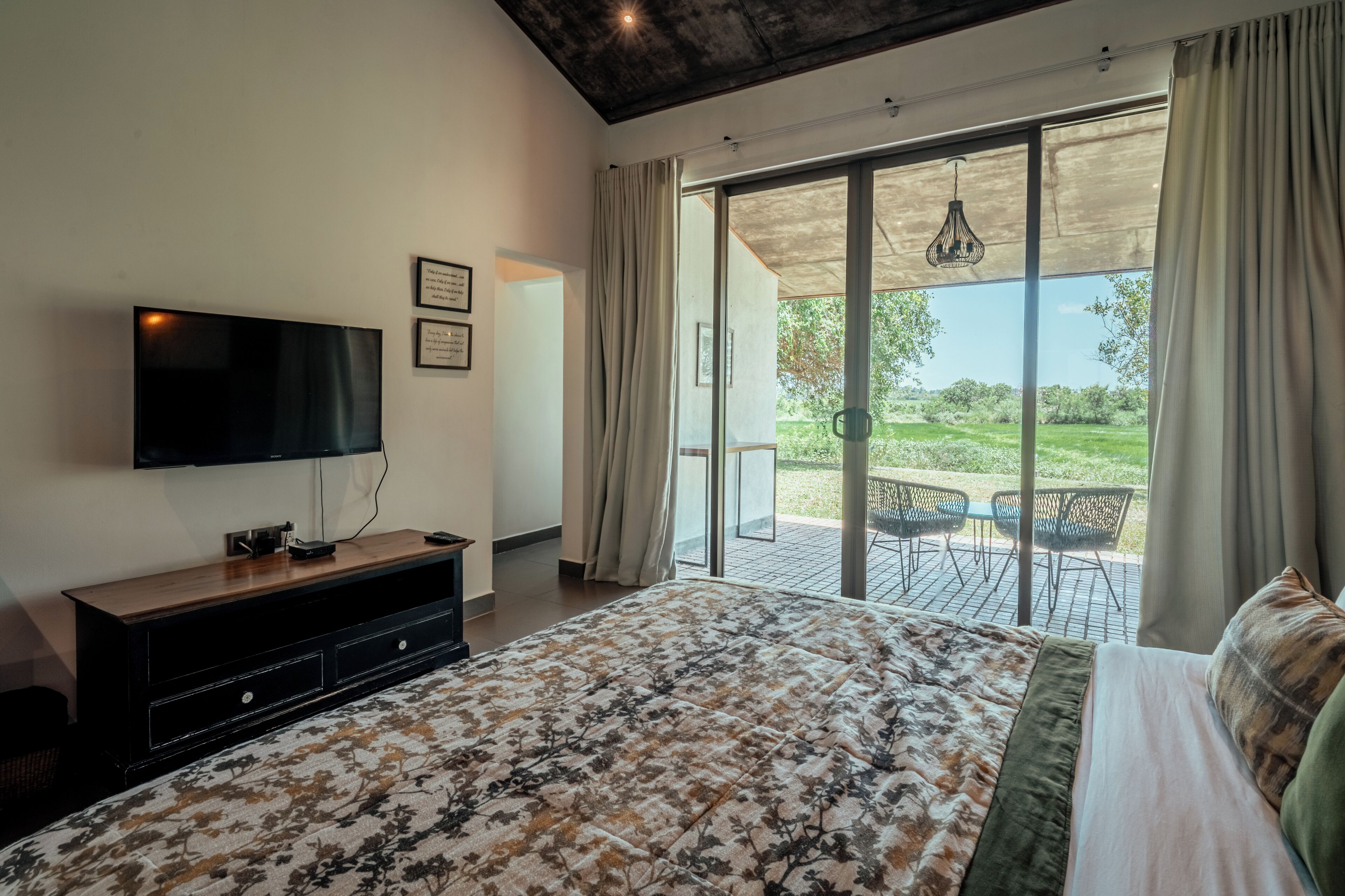 A bedroom with a bed, a TV, a wooden dresser, and a sliding glass door leading to an outdoor area with chairs.