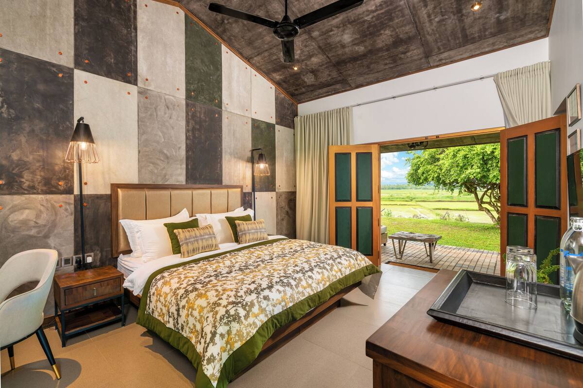 Un dormitorio con una cama grande, un escritorio, una silla y vistas a un paisaje verde a través de una puerta abierta.