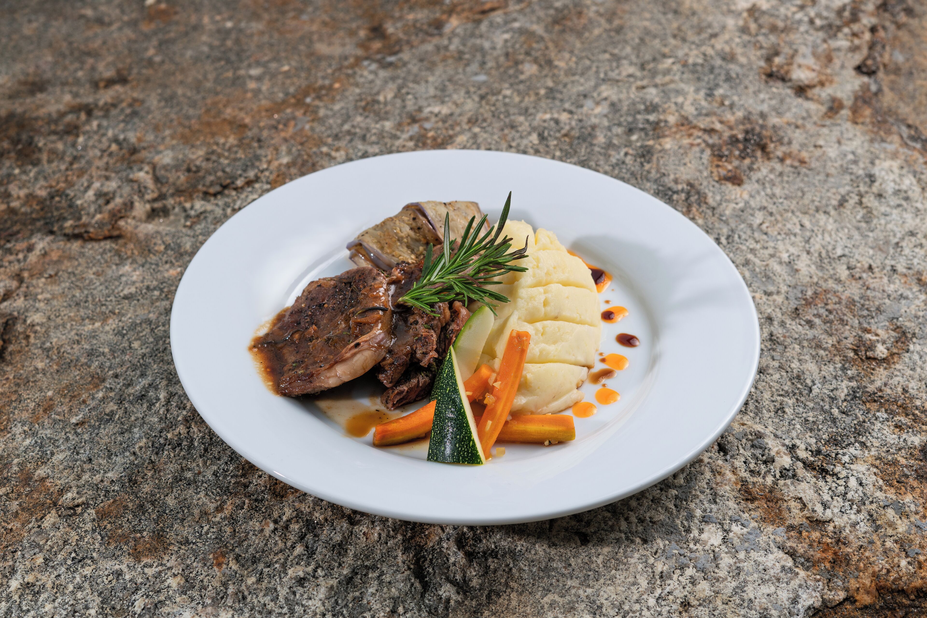 A plate of food with meat, mashed potatoes, and vegetables on a stone surface.