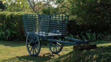 Un chariot en bois bleu, doté d’un dossier ajouré, est placé sur de l’herbe, avec un feuillage dense en arrière-plan.