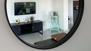 A modern living room with a large round mirror, a flat-screen TV, a teal chair, and a black entertainment unit. - 5 mn plages - 3 bedroom apartment - Downtown (Propriano)