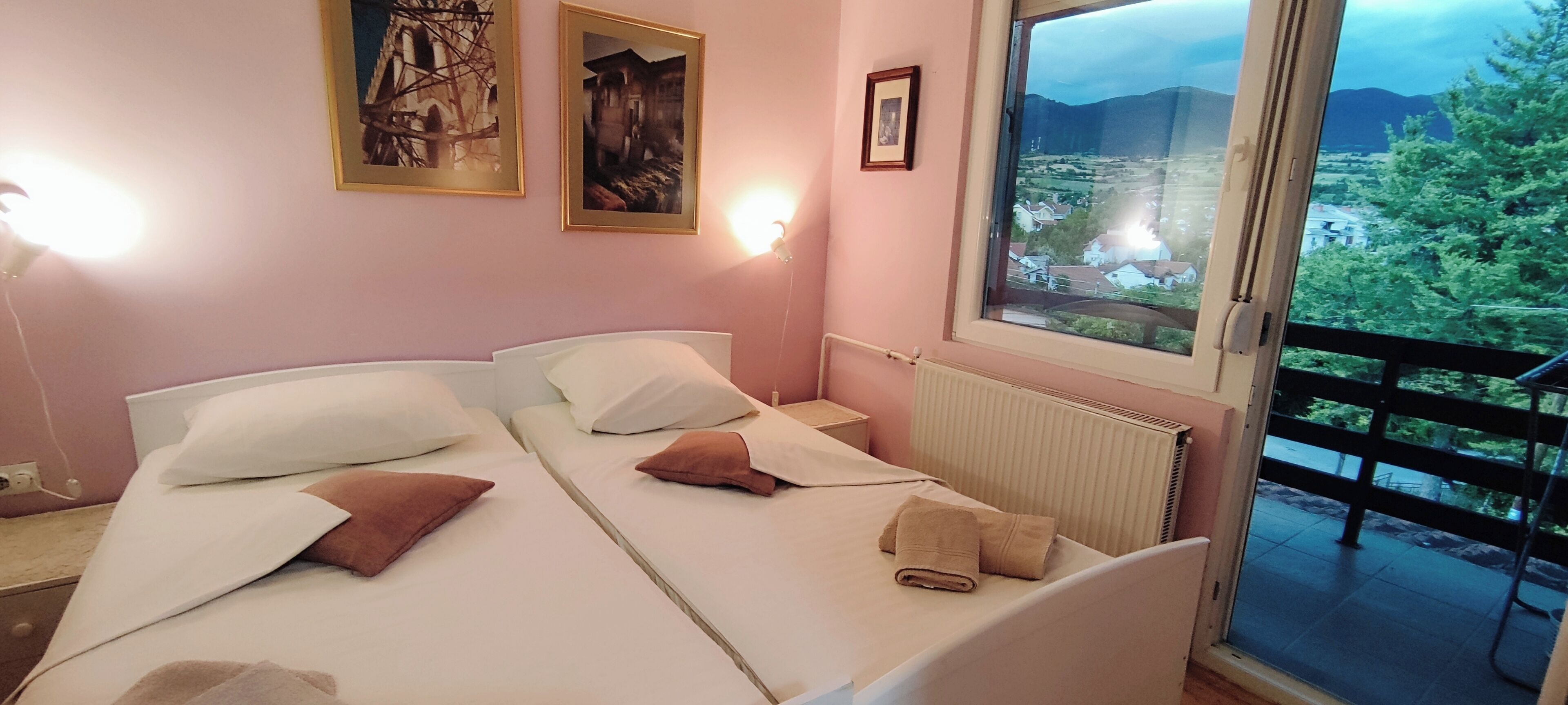 A bedroom with two beds, a window with a view of a village, and a radiator.