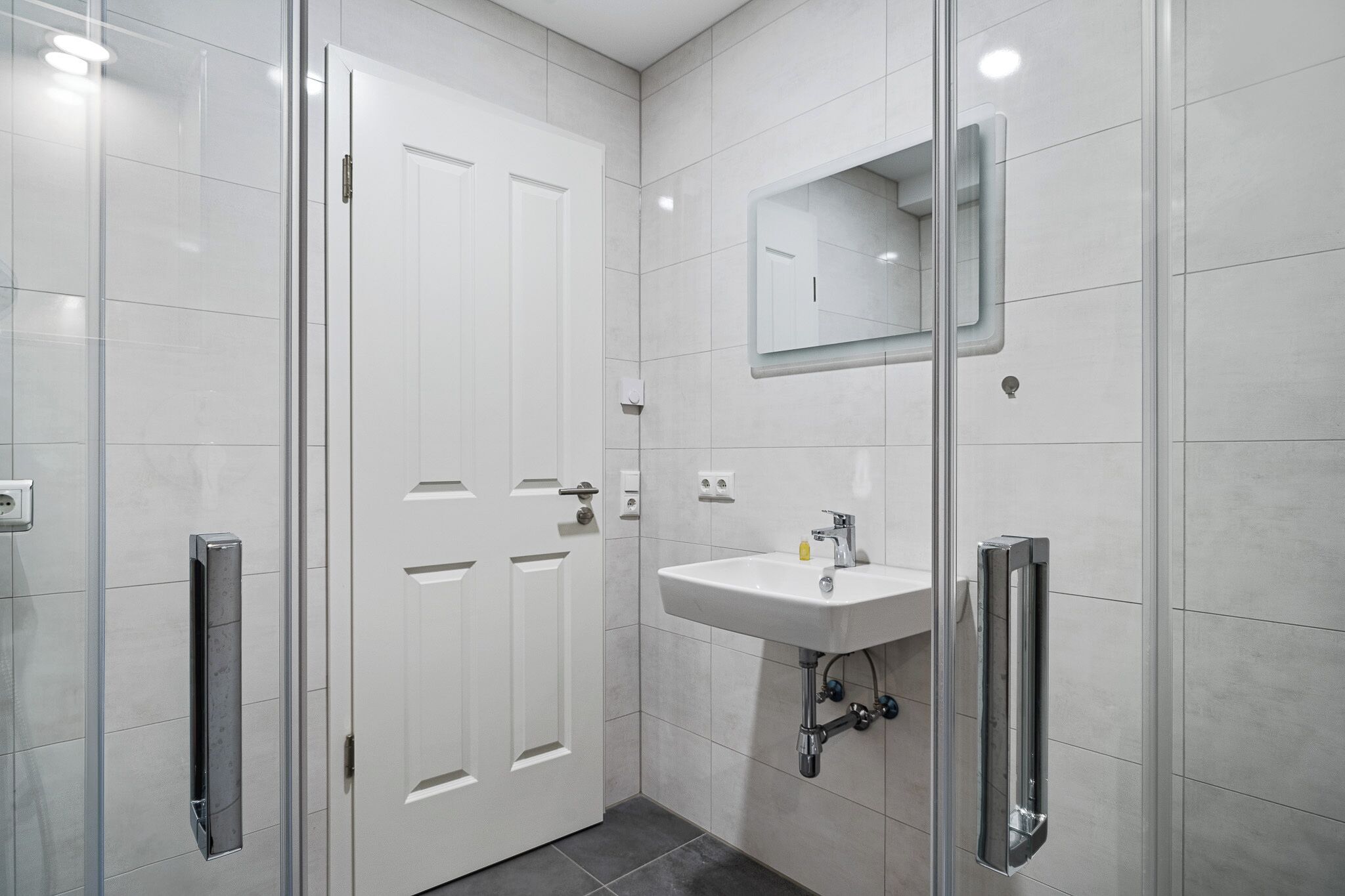 A modern bathroom with a glass shower enclosure, a white sink with a mirror above it, and a door with a handle.
