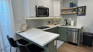 A modern kitchen with white cabinets, a built-in microwave, a stainless steel dishwasher, and a herringbone-patterned backsplash. There are two black bar stools and a small island with a white countertop.