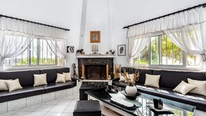 A modern living room with a fireplace, black leather sofas, a glass coffee table, and large windows with sheer curtains.