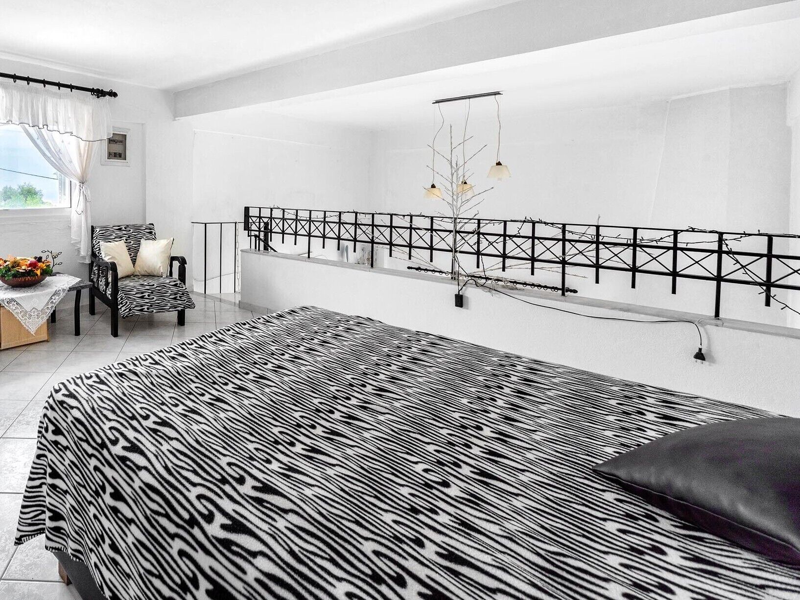 A modern bedroom with a zebra-patterned bedspread, a black and white chair, a small table with a vase of flowers, and a view of the outdoors through a window with sheer curtains.