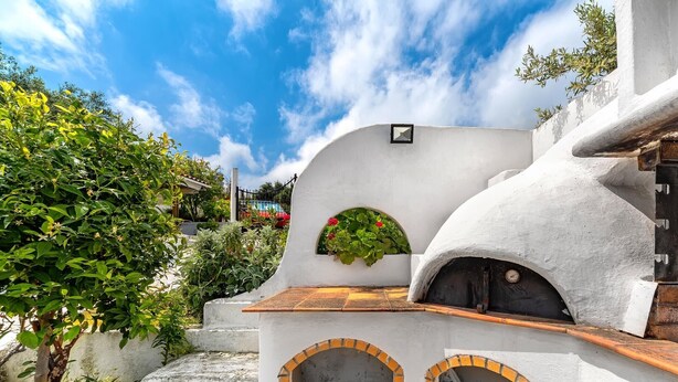 A white building with a tiled patio, a small garden, and a brick oven.